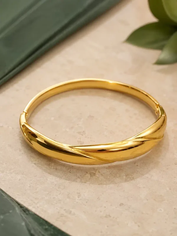 Polished gold bangle with overlapping twisted wave design styled on a neutral stone surface