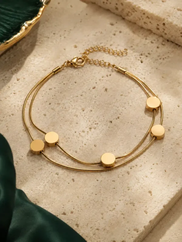 Double-strand gold-toned bracelet with five polished circular disc charms arranged on a neutral stone surface