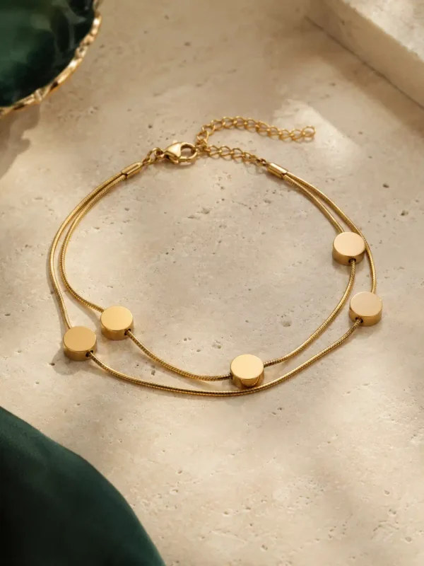 Adjustable gold-toned double-strand disc bracelet with lobster clasp and extension chain on a beige stone surface