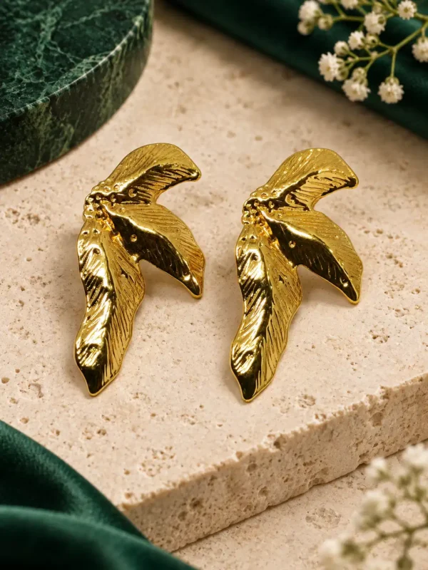 Pair of textured gold leaf and wing stud earrings styled on beige stone with green accents