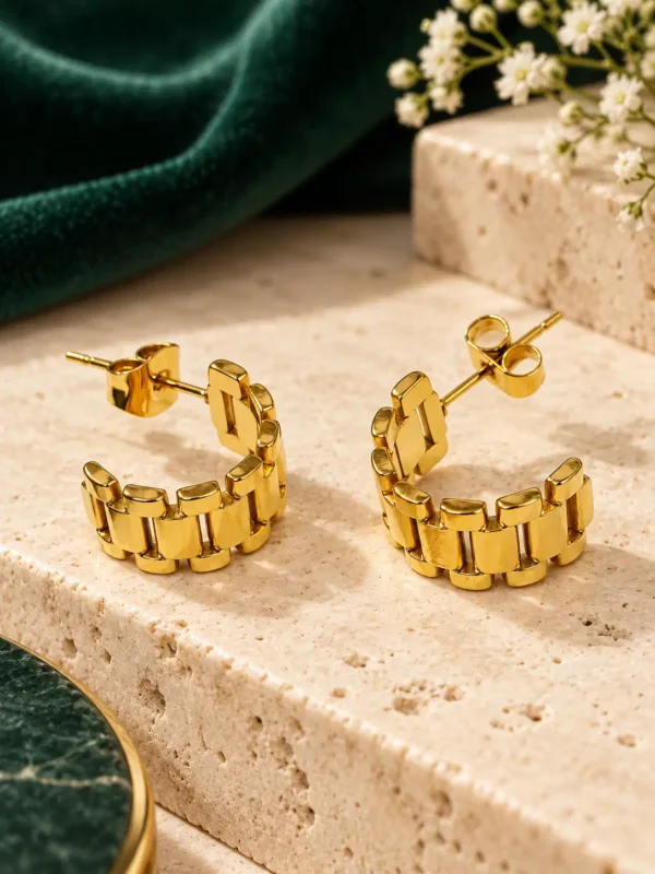 Pair of Elara gold watch-link hoop earrings on beige stone with green velvet backdrop