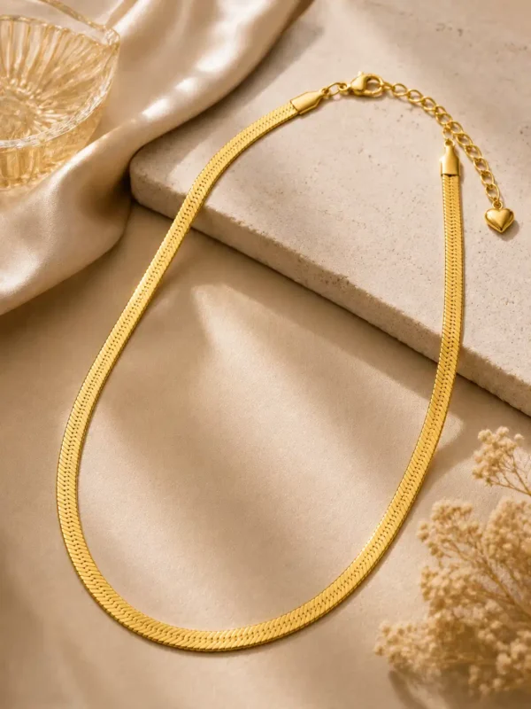 Gold-tone flat herringbone snake chain necklace with lobster clasp, extension chain and heart charm on a neutral flat lay.