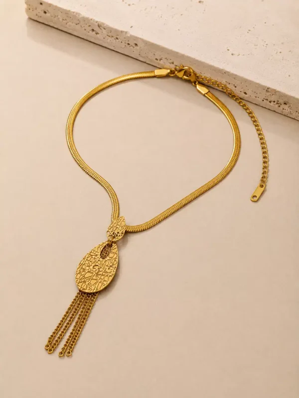 Macro product photograph of the Aurelia Textured Tassel Drop Necklace showing the detailed gold-tone pendant construction. A smaller leaf-inspired upper element leads into the elongated textured teardrop design with an inner opening and fine chain tassels, while the polished flat snake-style necklace is visible above.